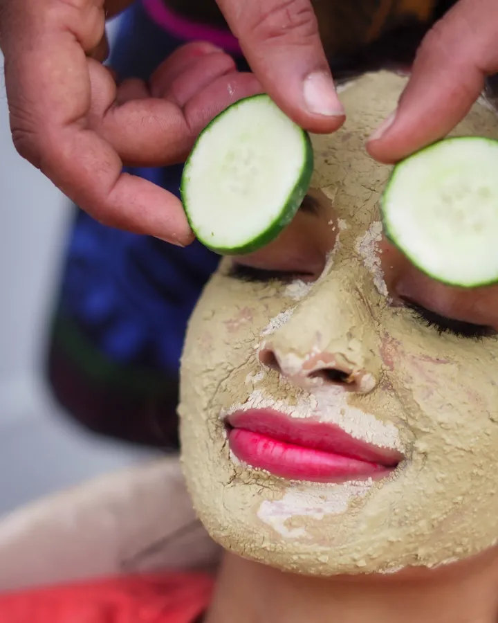 A woman lying back with a dried clay face pack and cucumber slices over her eyes