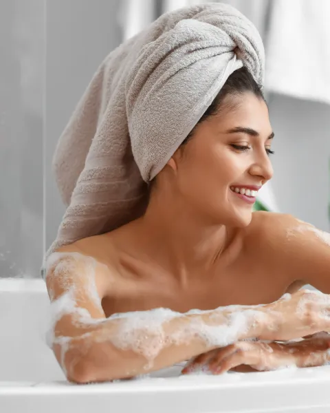 A woman in a hair towel in the bath, her arms lathered with soap