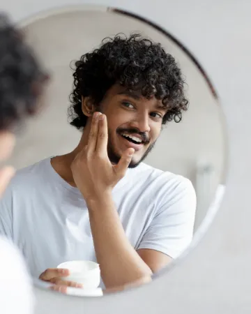 A man smiling in the mirror as he smooths cream into his cheek